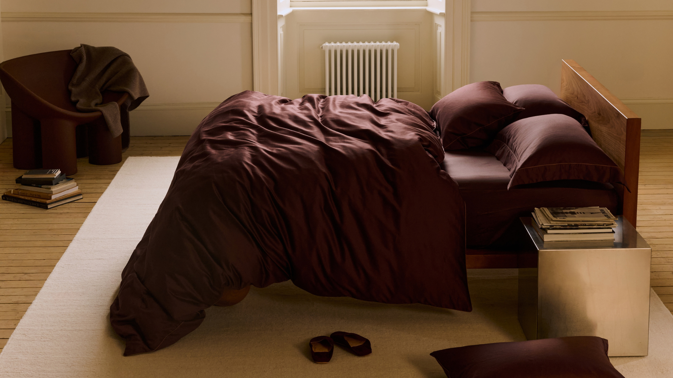 A deep brown-burgundy cotton sateen duvet set from Rise &amp; Fall styled on a bed in the middle of a white bedroom beside a steel side table