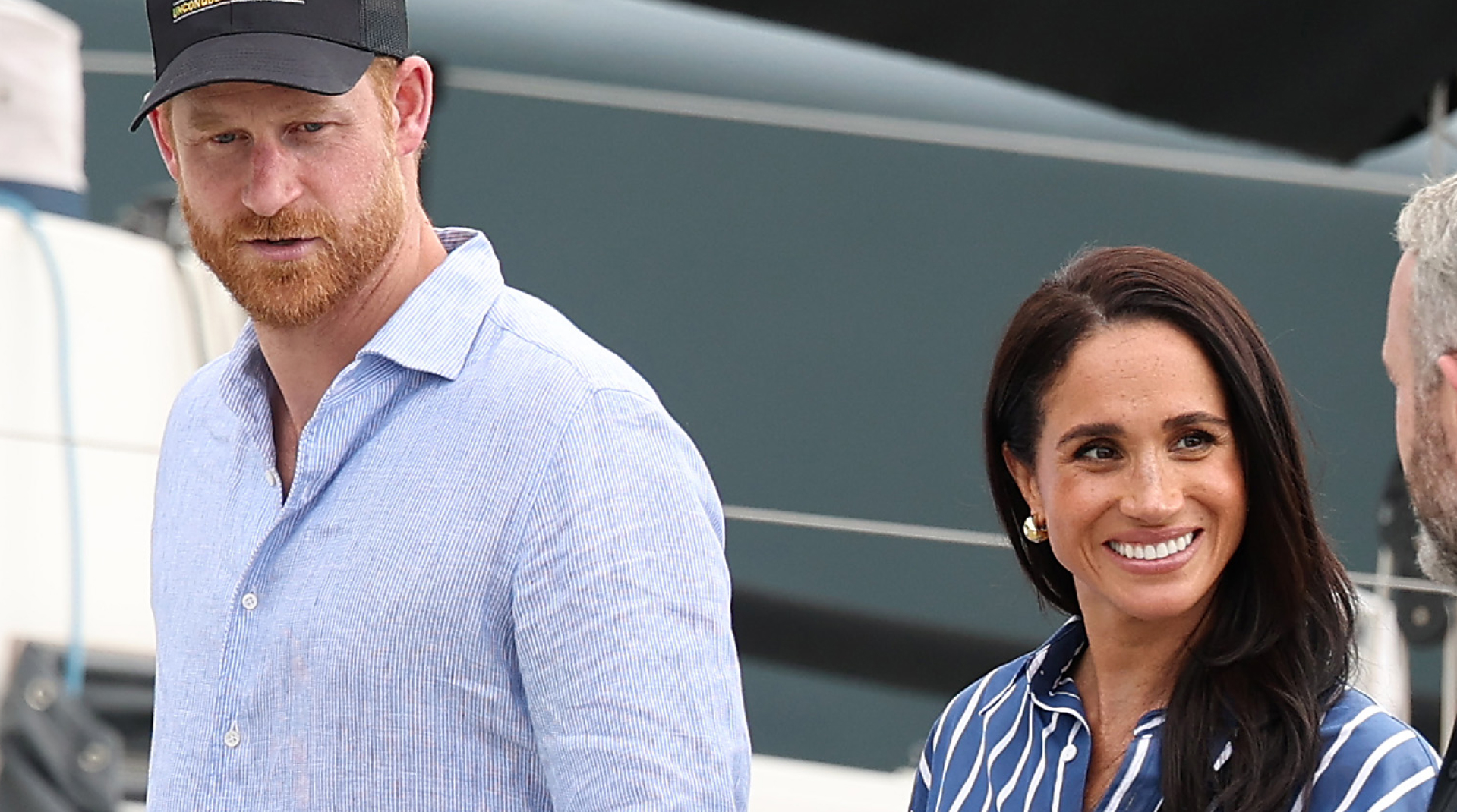 Prince Harry wears a baseball cap and a blue shirt, while Meghan Markle wears navy and white striped shirt and smiles