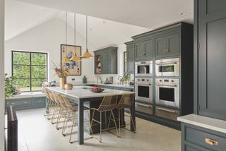 sage green kitchen with white walls and ceiling, stone floor, kitchen island with seating, brass pendant lights, downlighter, artwork, white countertop, window seat