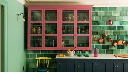 Dark blue lower kitchen cabinets and dusk pink upper glass cabinets. Green tiles are placed across the wall on one side, while the other side is painted green. A yellow and blue cushions chair sits beneath the upper cabinets.
