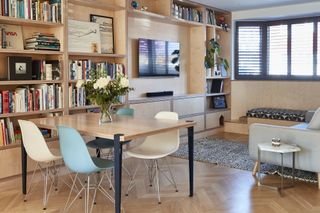 A plywood living room with built-in storage, leading into an open plan dining area with Eames-style chairs