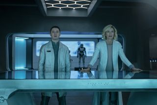 Adun (Ryan McParland) and Dr Catherine Halsey (Natascha McElhone) stand in her laboratory, and Dr Halsey is resting her hands on the large steel desk in front of her. They are both looking at an unseen object behind the camera - his expression is slightly concerned, hers is one of rapt fascination. There is an empty suit of Spartan armour on a plinth behind them in the background. The laboratory itself is very sleek, minimalist, with steel fixtures and a tessellated hexagon pattern above them in the centre of the ceiling.