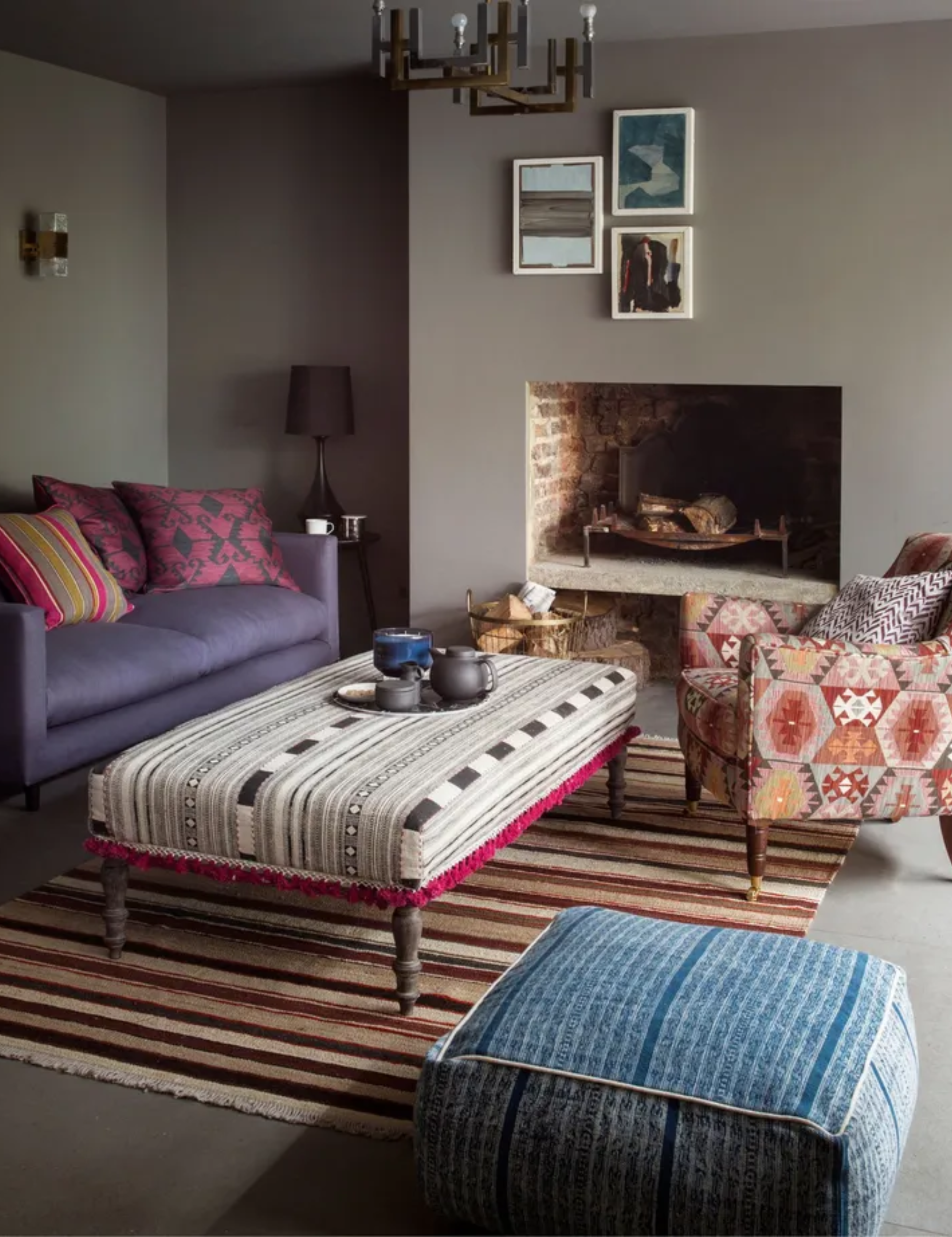 A neutral brown living room with contrasting patterned ottoman, chair and pouffe, striped brown rug and purple sofa with bright pink cushions and three framed pictures in white frames below a metal industrial pendant light