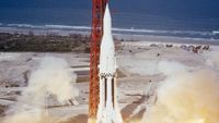 a white rocket with black vertical stripes on its first stage lignites its engines next to a red support tower near the seashore