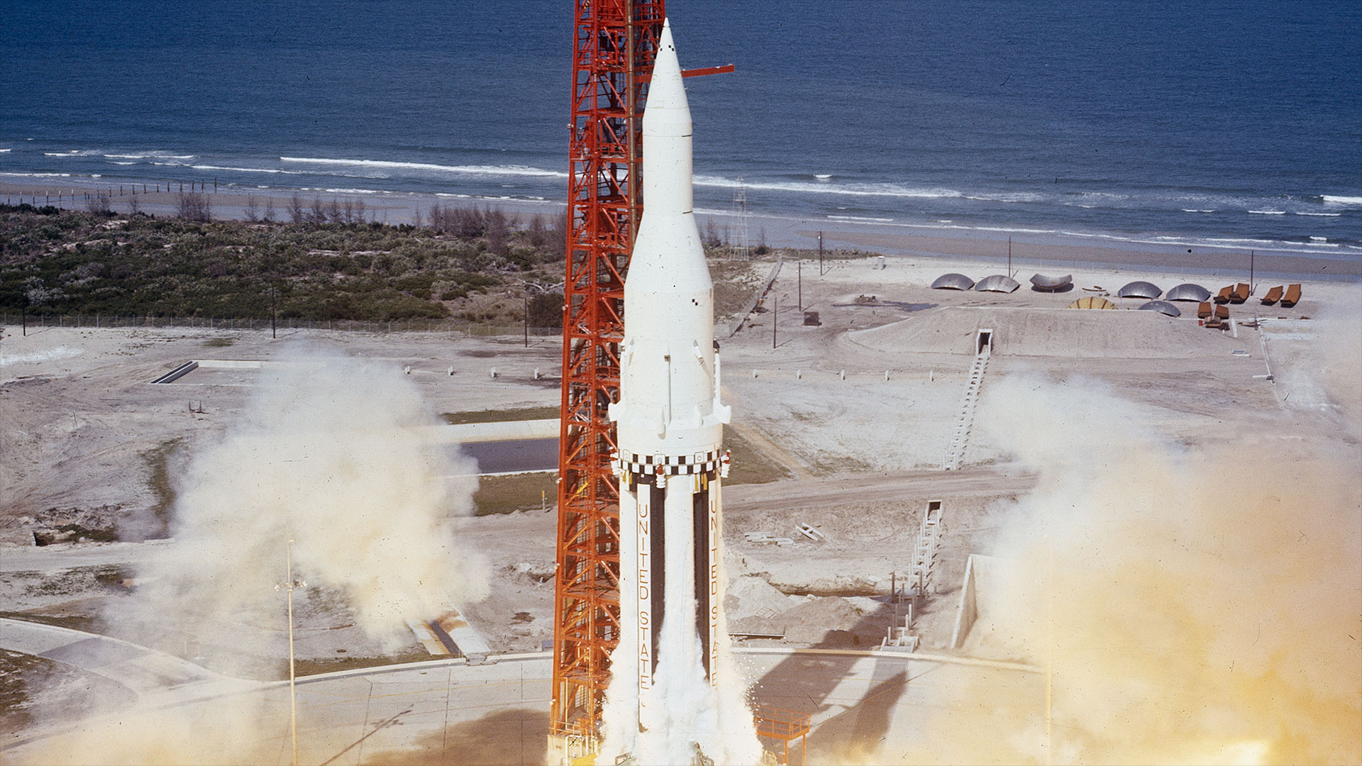a white rocket with black vertical stripes on its first stage lignites its engines next to a red support tower near the seashore