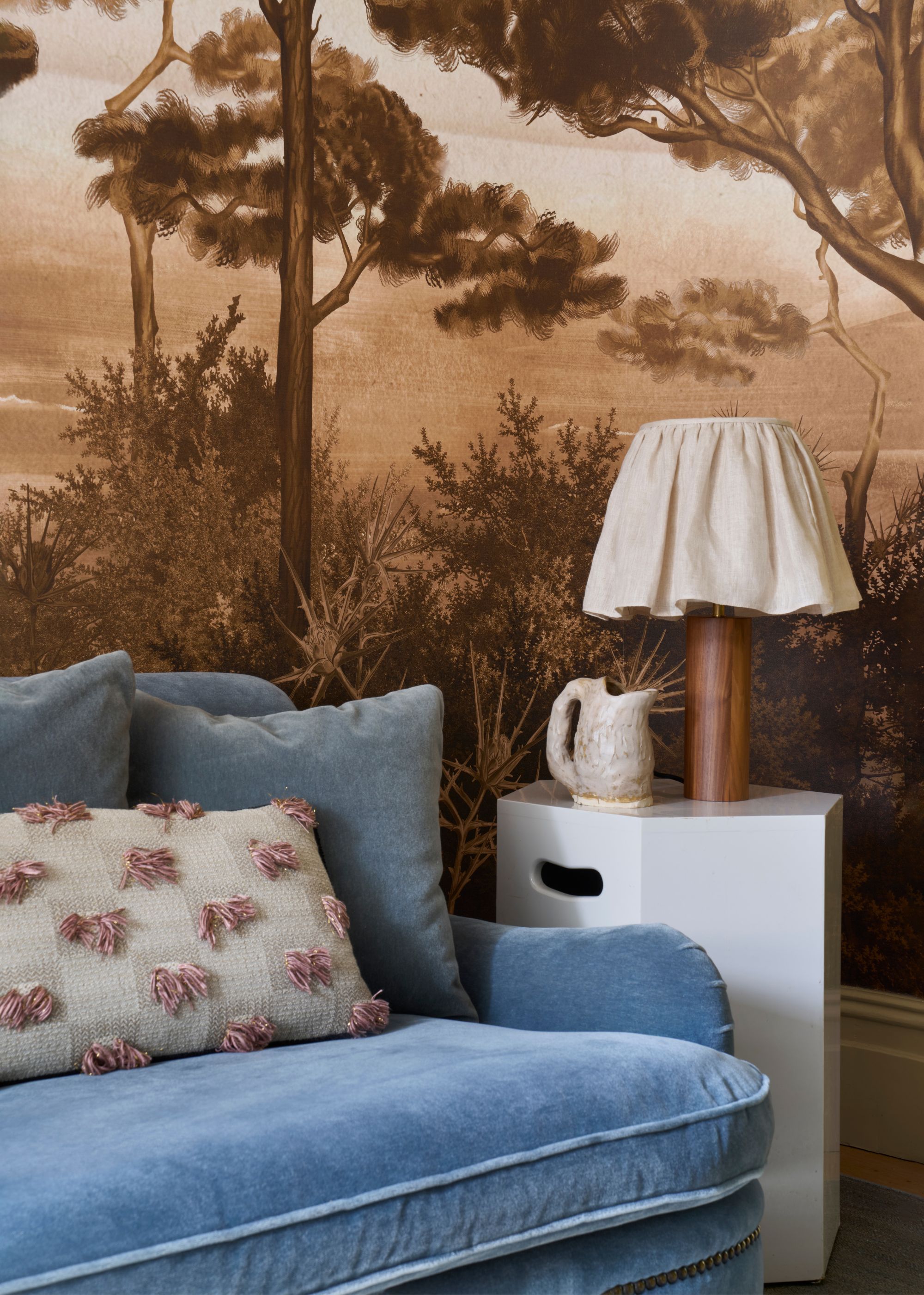 The corner of a living room with a side table and a lamp on top of it, as well as a decor piece on the table. There is a view of the corner of a light blue vintage sofa with a cushion on top.