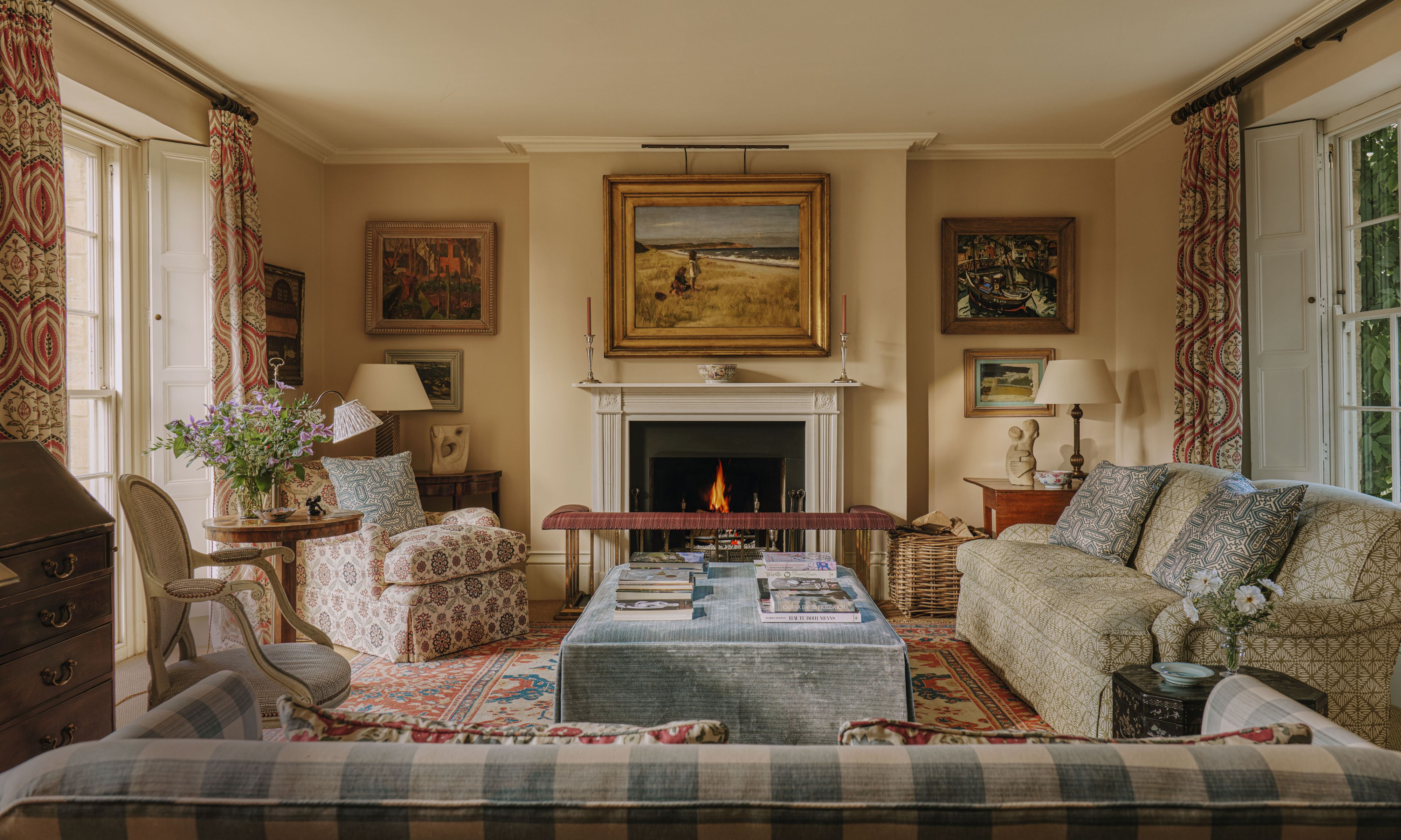 Cozy traditional living room featuring an open fireplace, light blue ottoman, framed artwork, and patterned sofas on a vintage rug.