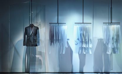 View of the Kilgour ‘All the Work’ installation featuring a dark suit jacket on a half mannequin hanging from a black bar behind glass. Beside it is three rails of paper drafting patterns on hangers