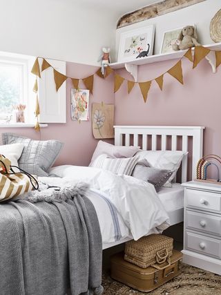 pink girls bedroom with gray blanket and pillows, baskets, old suitcase, bunting, shelf above bed, white bed and side table