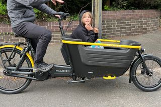 The Raleigh Stride 2 e-cargo bike with a child in the front cargo tub with a brick wall behind