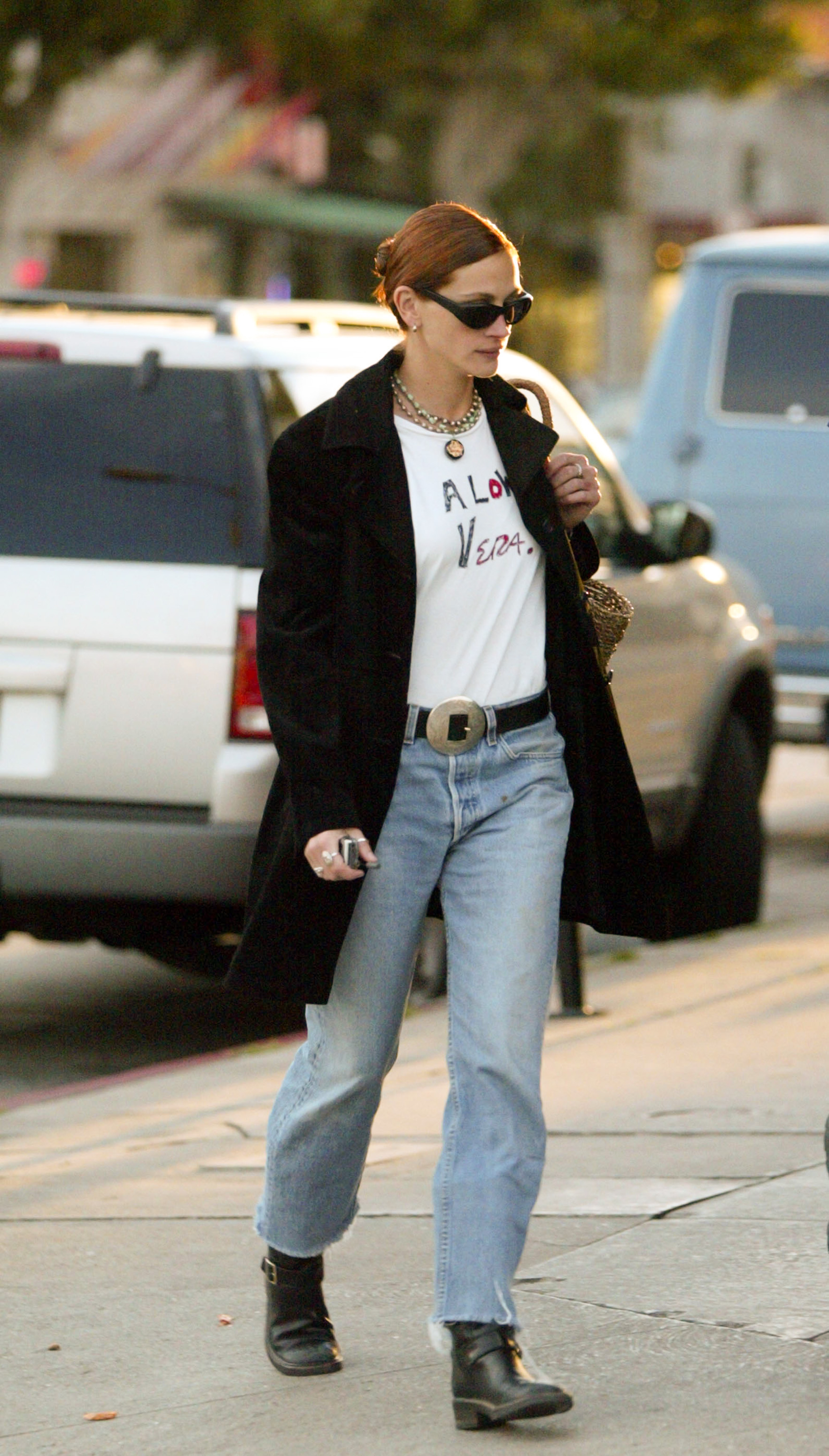 An image of actor Julia Roberts from the '90s walking outside wearing a black coat, a graphic T-shirt that says &amp;quot;A Low Vera.&amp;quot;, light-wash jeans and black boots.