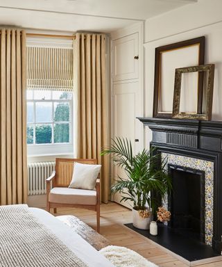 A bedroom with tall, thick, taupe drapes, a black ornate fireplace with blue and yellow floral decals, a potted Kentia palm and rattan accent chair