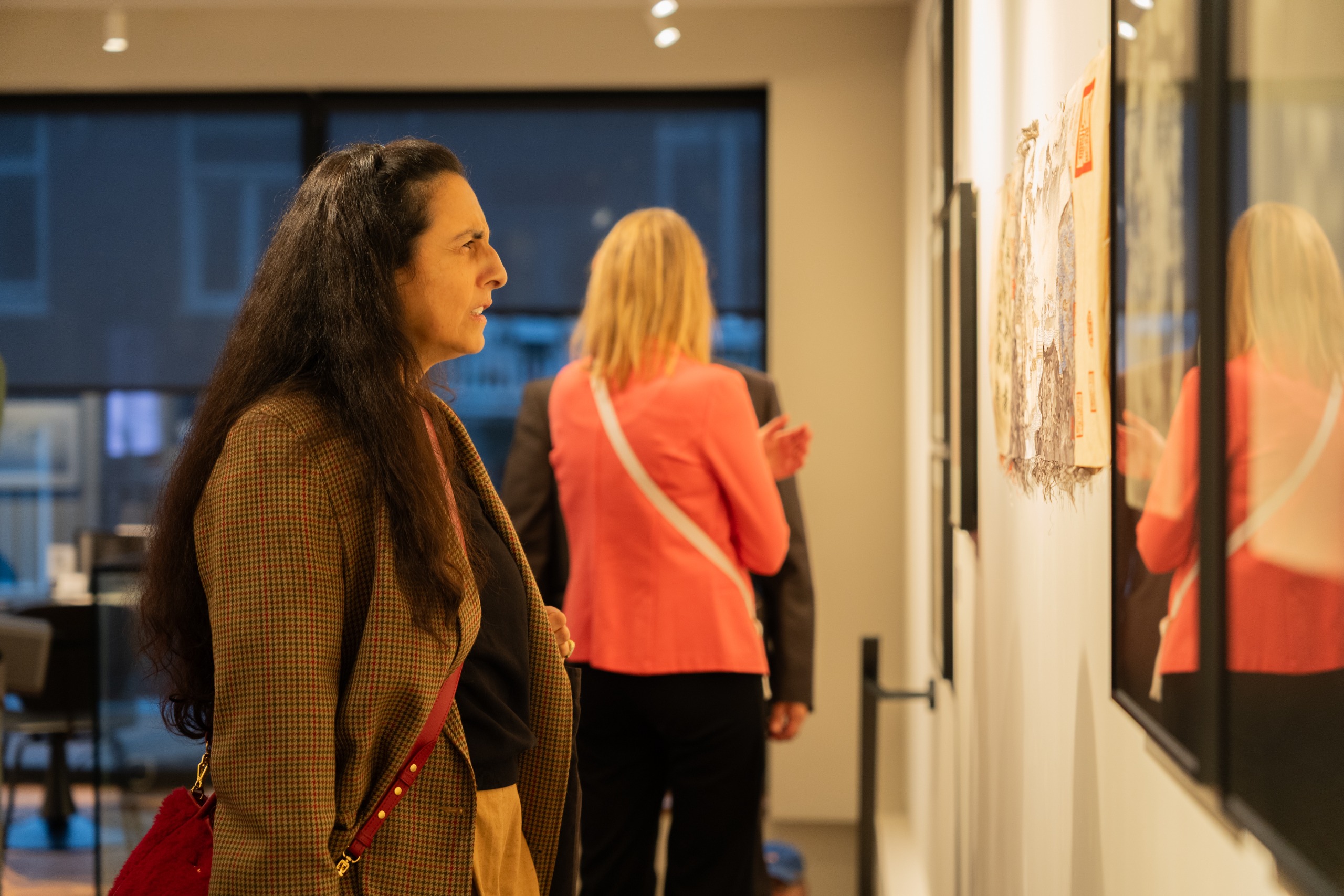 A woman in a houndstooth jacket studies a delicate, finely detailed silk work on the gallery wall, while other visitors talk in the background.