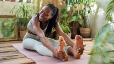A woman performs a forward fold on an exercise mat. She is sitting with her legs extended and bending forward to grasp her feet. 