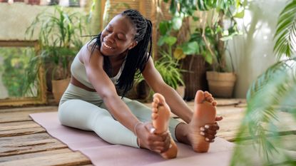A woman performs a forward fold on an exercise mat. She is sitting with her legs extended and bending forward to grasp her feet.