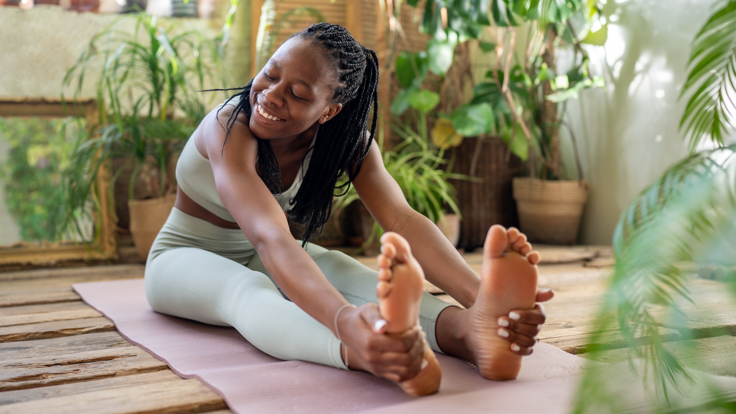 A woman performs a forward fold on an exercise mat. She is sitting with her legs extended and bending forward to grasp her feet. 