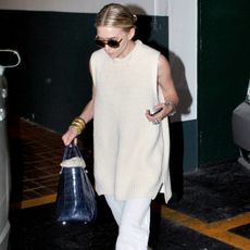 Ashley Olsen walking in a car garage in Los Angeles in chic summer look with a slicked-back bun, round sunglasses, a ribbed sleeveless tunic sweater, Sidney Garber gold bracelets, a blue croc The Row bag, and white linen pants