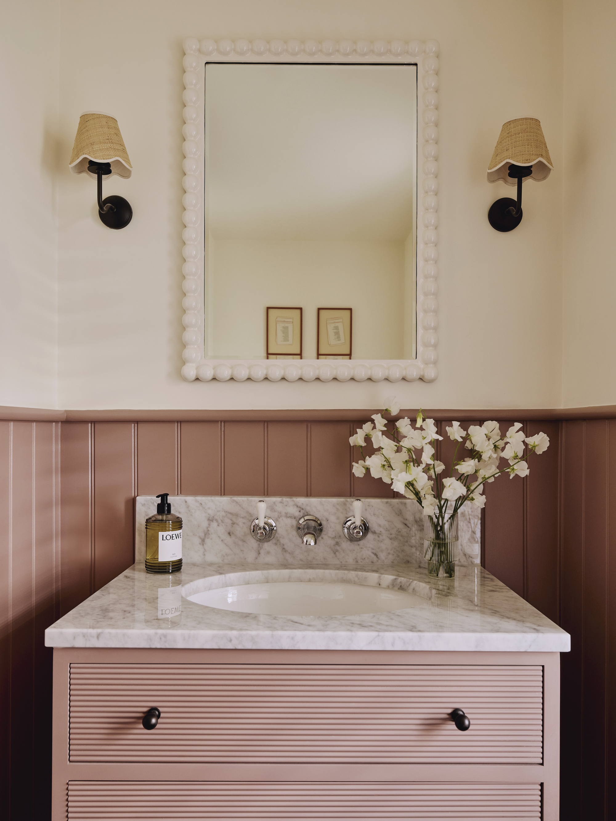 bathroom with cream walls, wall sconces on either side of a white bobbin mirror, with dark pink panelling, with a freestanding vanity with marble countertop and pink drawers