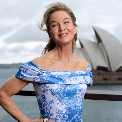 Renee Zellweger wearing an off-the-shoulder blue and white dress. She also is wearing a half up half down hairstyle and smiling at the camera with her hand on her hip.
