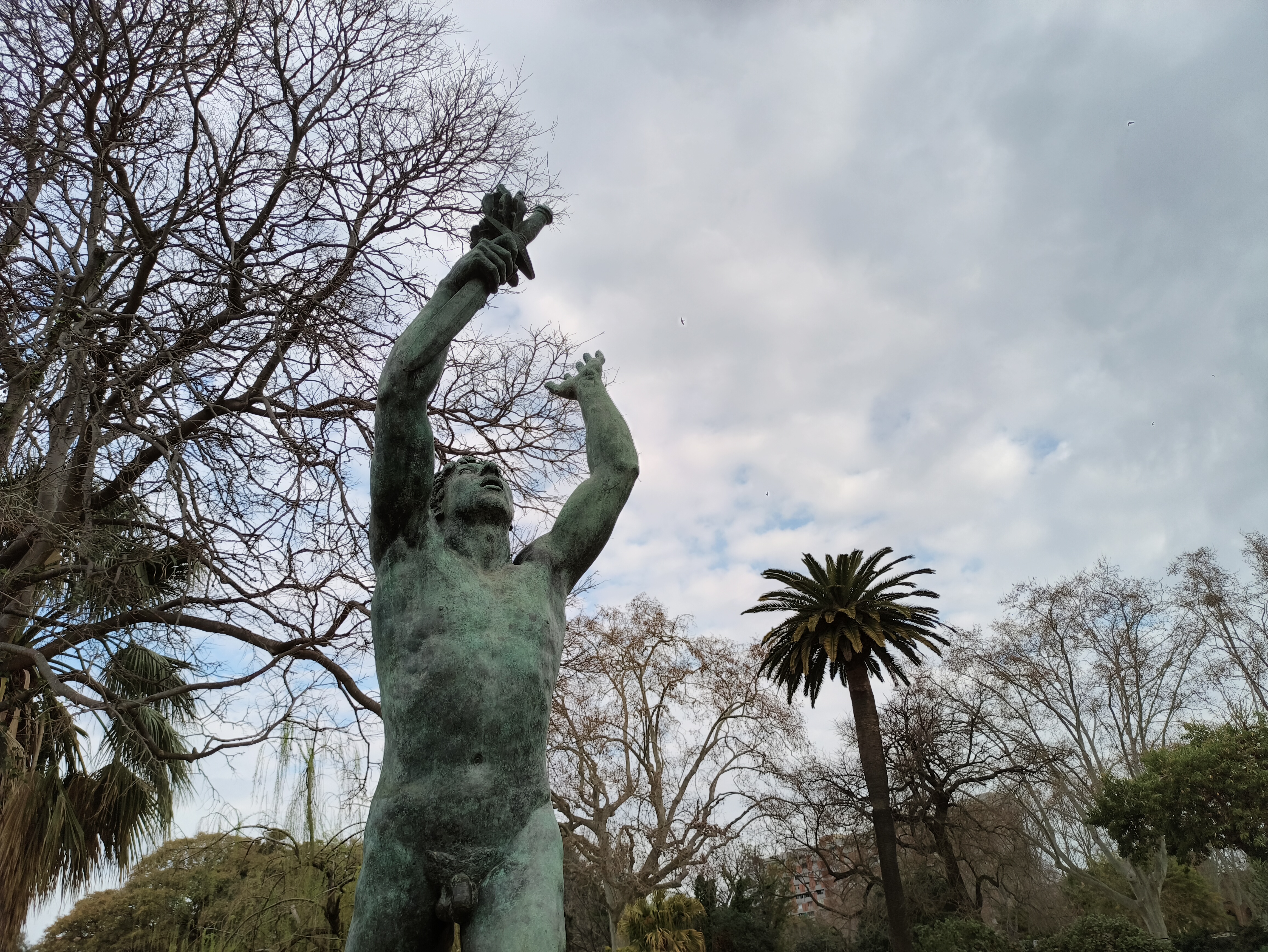 A bronze statue of a muscular man reaching upward toward a cloudy sky is framed by the branches of a large leafless tree and a single tall palm tree.