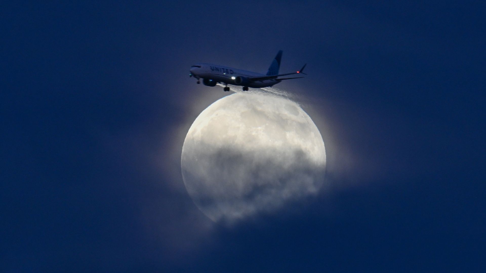 A commercial jet is photographed as it appears to touch the edge of a full moon in a cloudy sky.