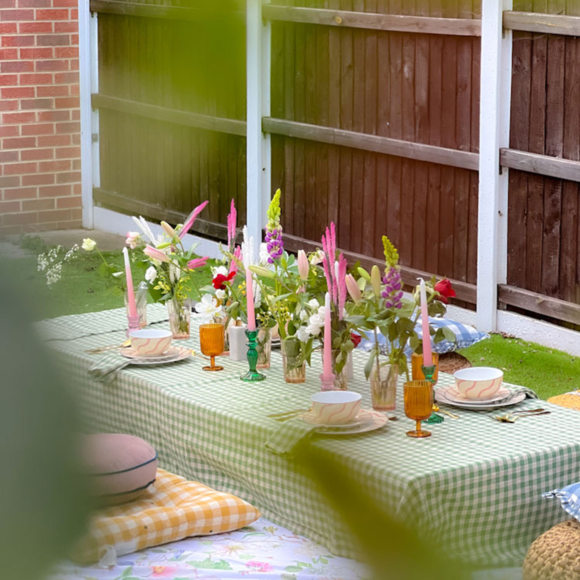 Outdoor table with green and white gingham tablecloth, flowers in tumblers and flatware with pink and white squiggle pattern alongside yellow and white gingham floor cushion and round pink pillow