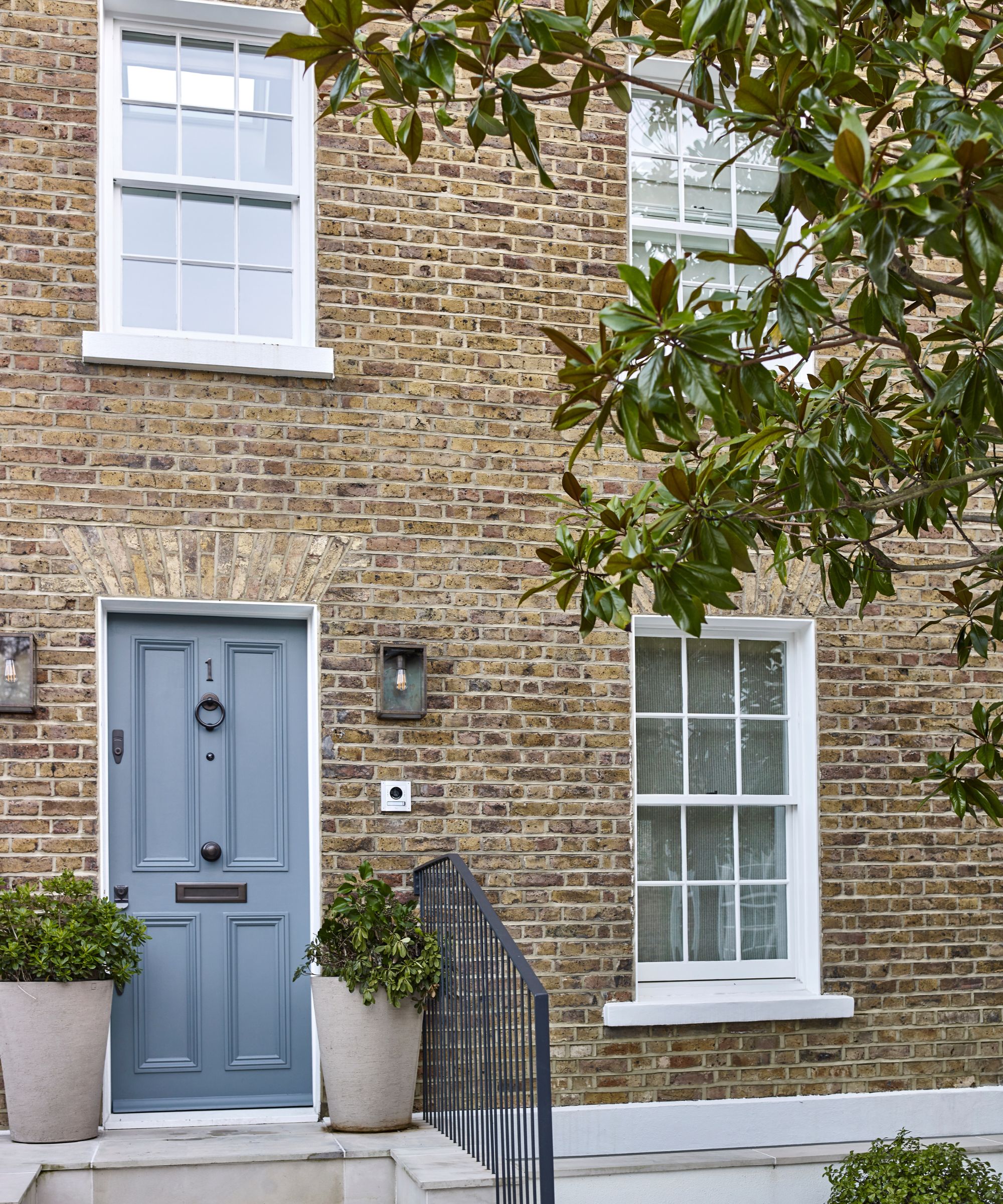 Brick townhouse with large white windows, a grey painted front door with a knocker, two potted plants on either side, sconces on the wall, and steps leading up with iron railings.