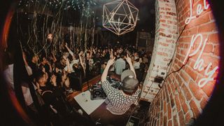 The interior of a nightclub with a sultry lounge and DJ getting the crowd amped up with arms in the air. 