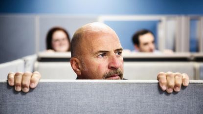 A man peers over the top of a cubicle at work.
