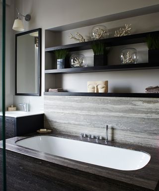 A gray bathroom scheme with a sunken bath tub below black wall shelves displaying sculptures and towels