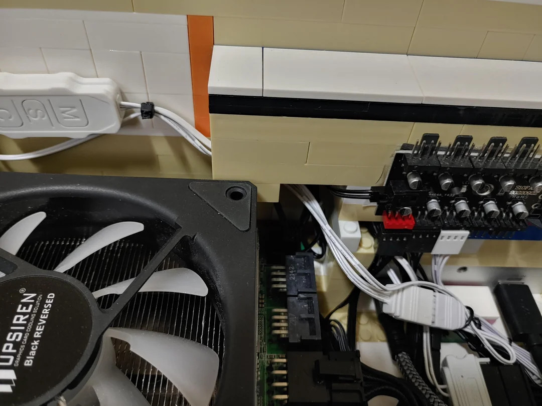 Close-up of a computer setup with visible fan, wires, and circuit board. Beige and orange LEGO bricks enclose the components, creating a playful tone.