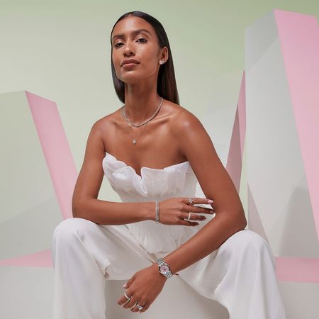 Model wearing Goldsmiths jewellery and a watch infront of a pink and green pastel background