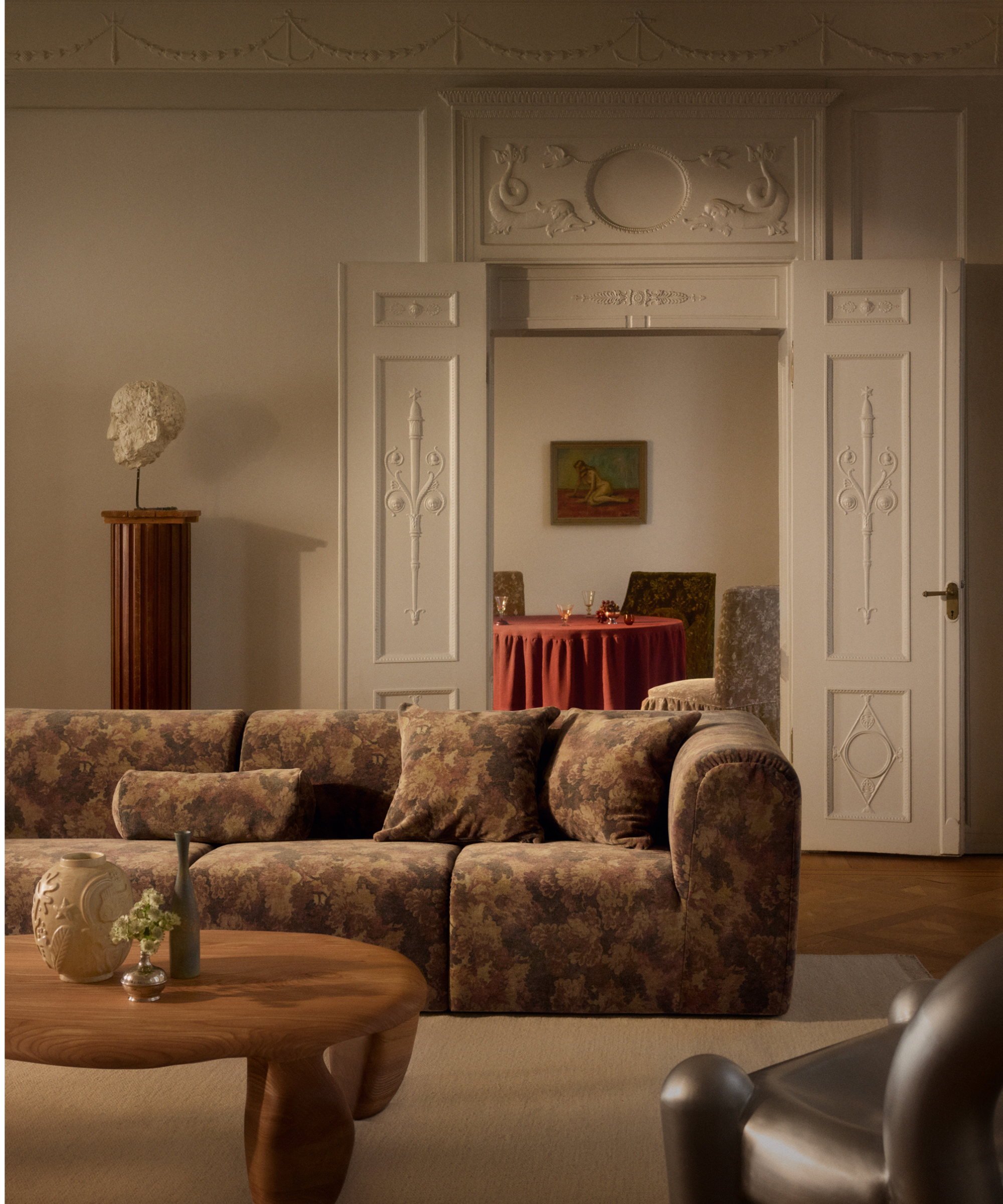 Tapestry-patterned sofa and matching throw pillows styled in an opulent living room with ornate moulding, a Grecian bust, and low-slung, organically-shaped coffee table.