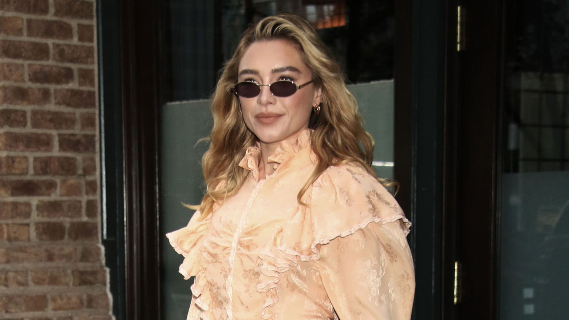 Florence Pugh steps out in New York wearing white flared trousers over heels. She styles her trousers in a peach frill blouse and carries a woven top handle bag.
