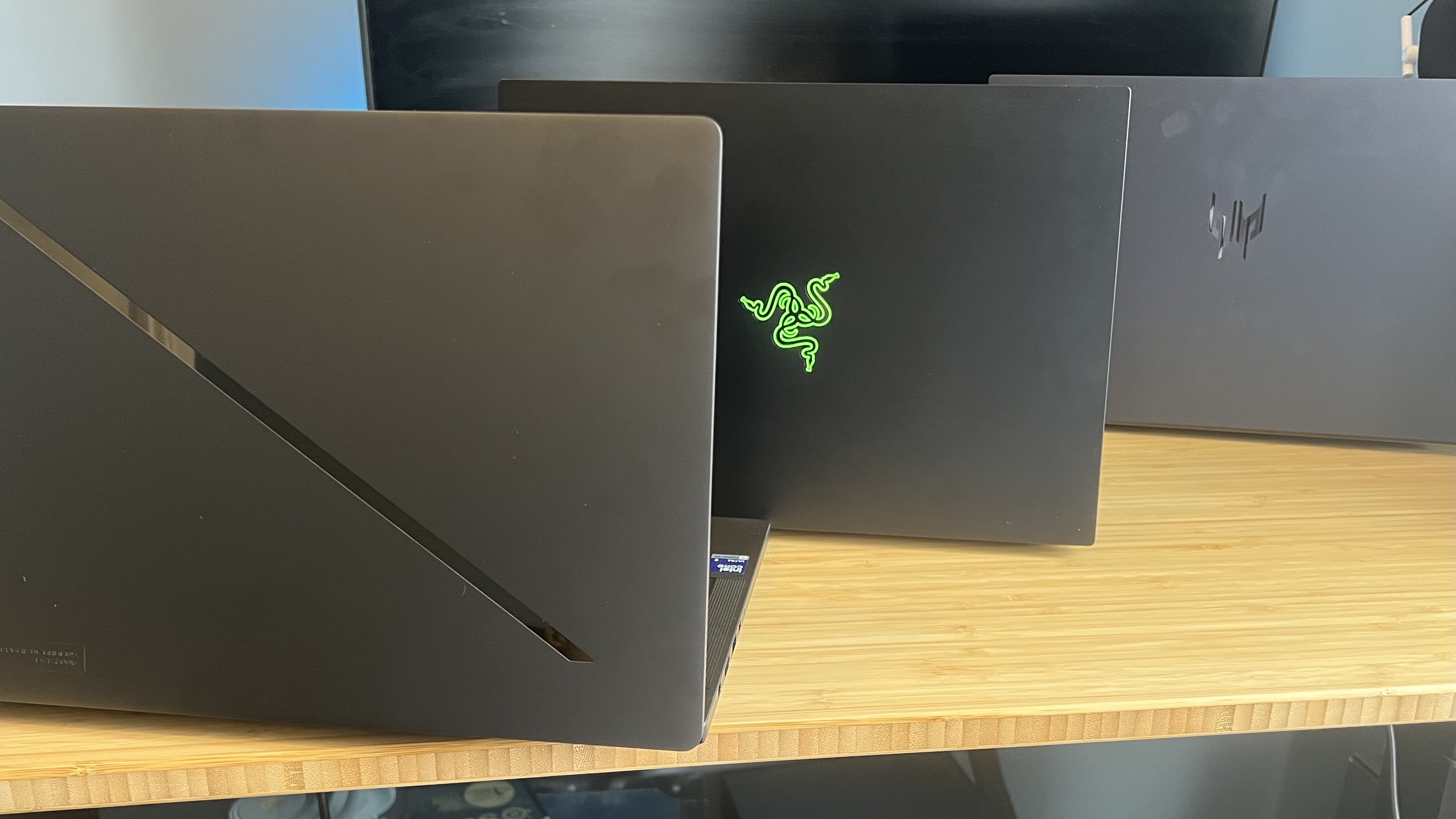 Asus ROG Zephyrus G14, Razer Blade 14, and Acer Predator Triton 14 AI gaming laptops lined up on a wooden desk