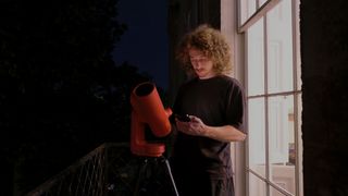 A man with long hair stood next to a bright window and red smart telescope at night whilst looking at a smartphone.