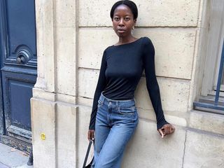 style influencer Sylvie Mus poses on a Paris sidewalk with a statement door wearing a black boatneck top, high-waisted jeans, and a minimal black bag