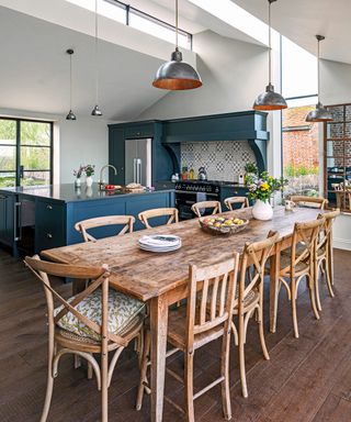 Open plan kitchen diner with modern white ceiling with skylights, large wooden dining table, dark wooden flooring, blue kitchen cabinetry, blue kitchen island with black countertop, low hanging metal pendant lights over table and island