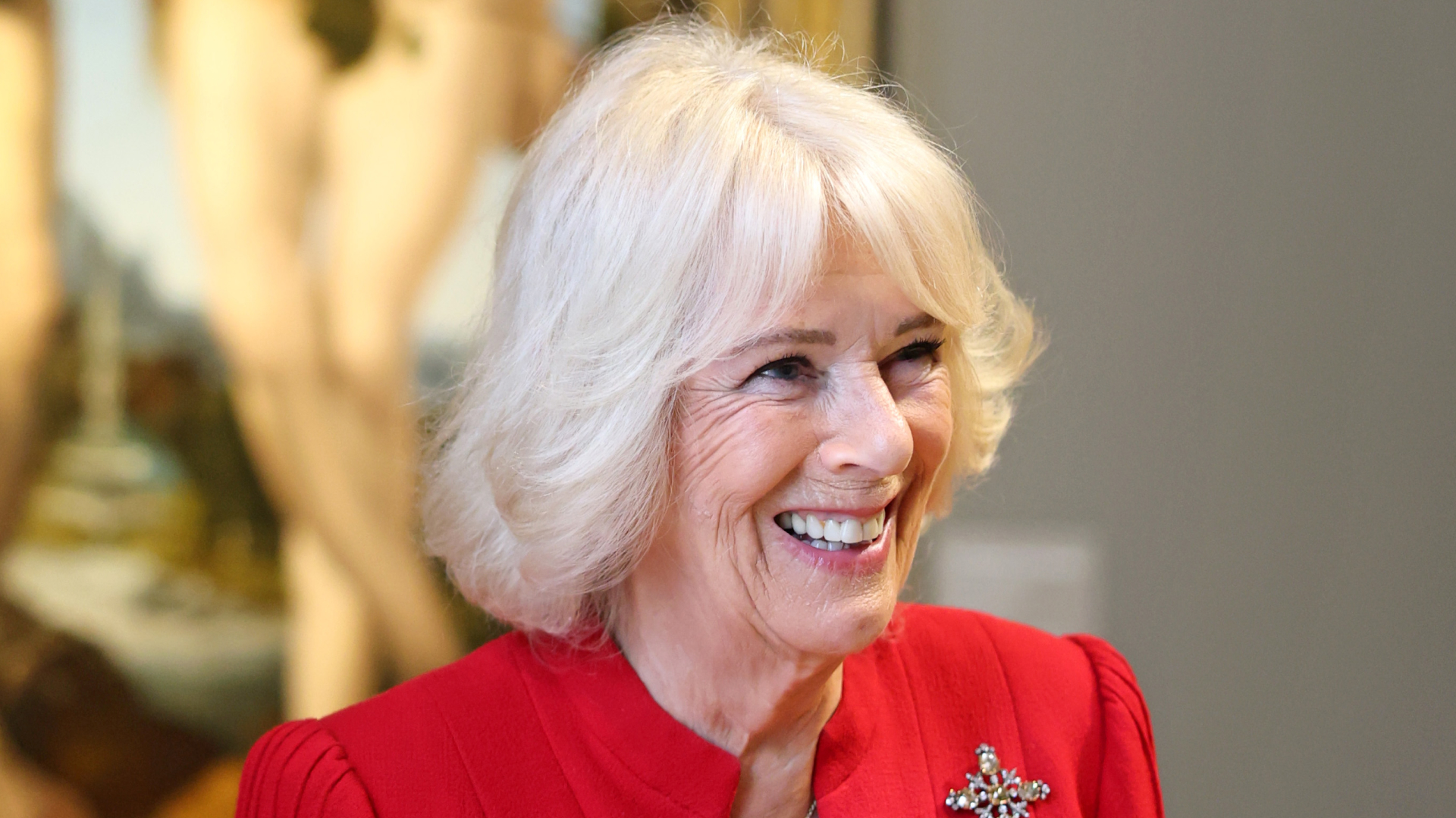 Queen Camilla wearing a red dress and diamond brooch smiling