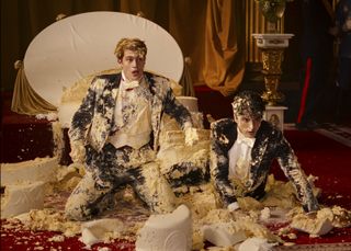 Henry (Nicholas Galitzine) and Alex (Taylor Zakhar Perez) are wearing their wedding finery, absolutely covered from head to toe in cake and frosting. A collapsed table and the remnants of the cake lie behind them. Both of them look shocked and embarrassed.