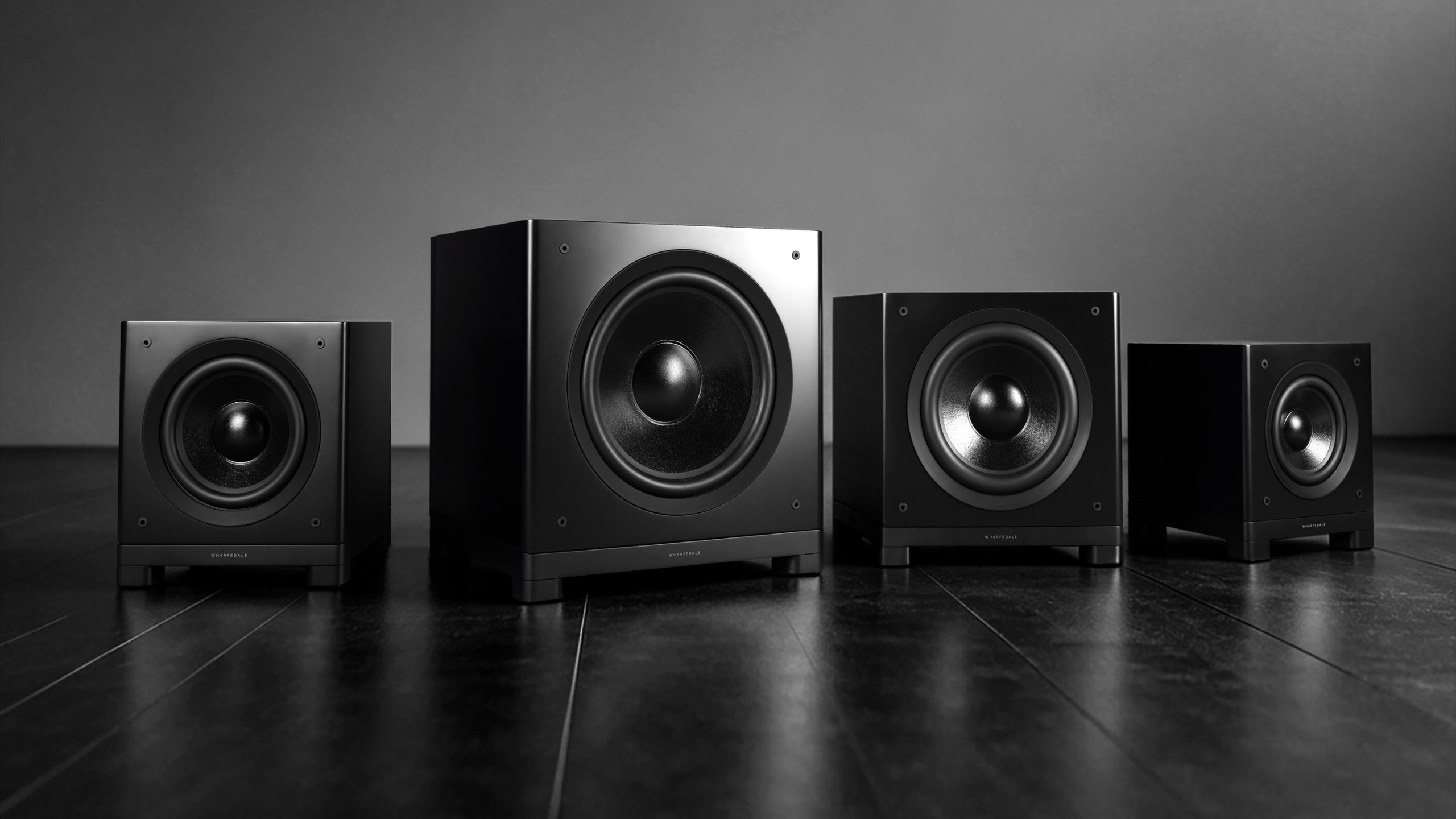 Four black Wharfedale Nova Series subwoofers arranged in a row on a black surface.