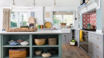 A kitchen with blue cabinets, white walls, pink and white tiles, central island and wooden floors, shelves full of cookware and baking tools