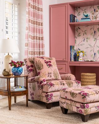 Corner of a room with pink patterned accent chair, matching foot rest, pink shelves and patterned pink wallpaper and pink curtains