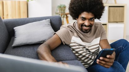 A young man smiles as he looks at his laptop screen and his smartphone in his living room.