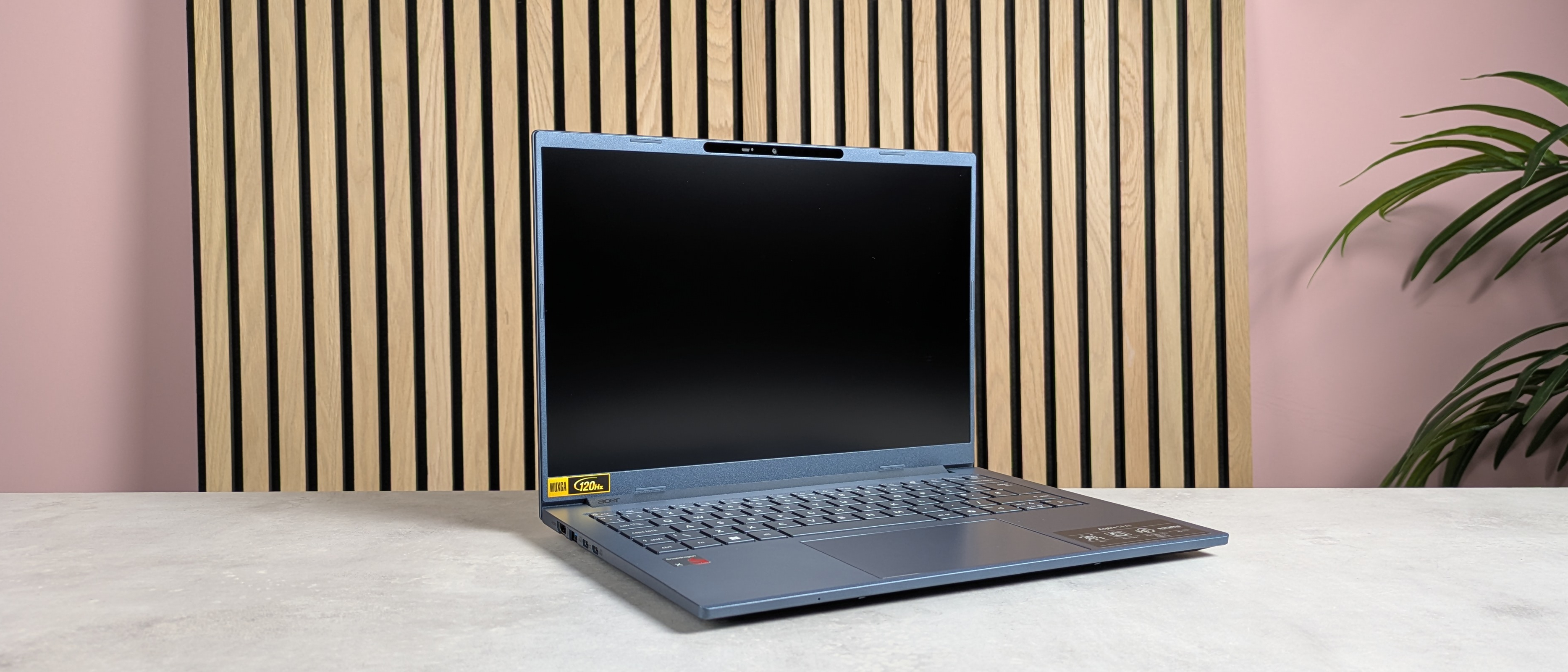 Three-quarter view of Acer Aspire 14 AI (2025) open on desk with wooden panel, plant, and pink wall in background