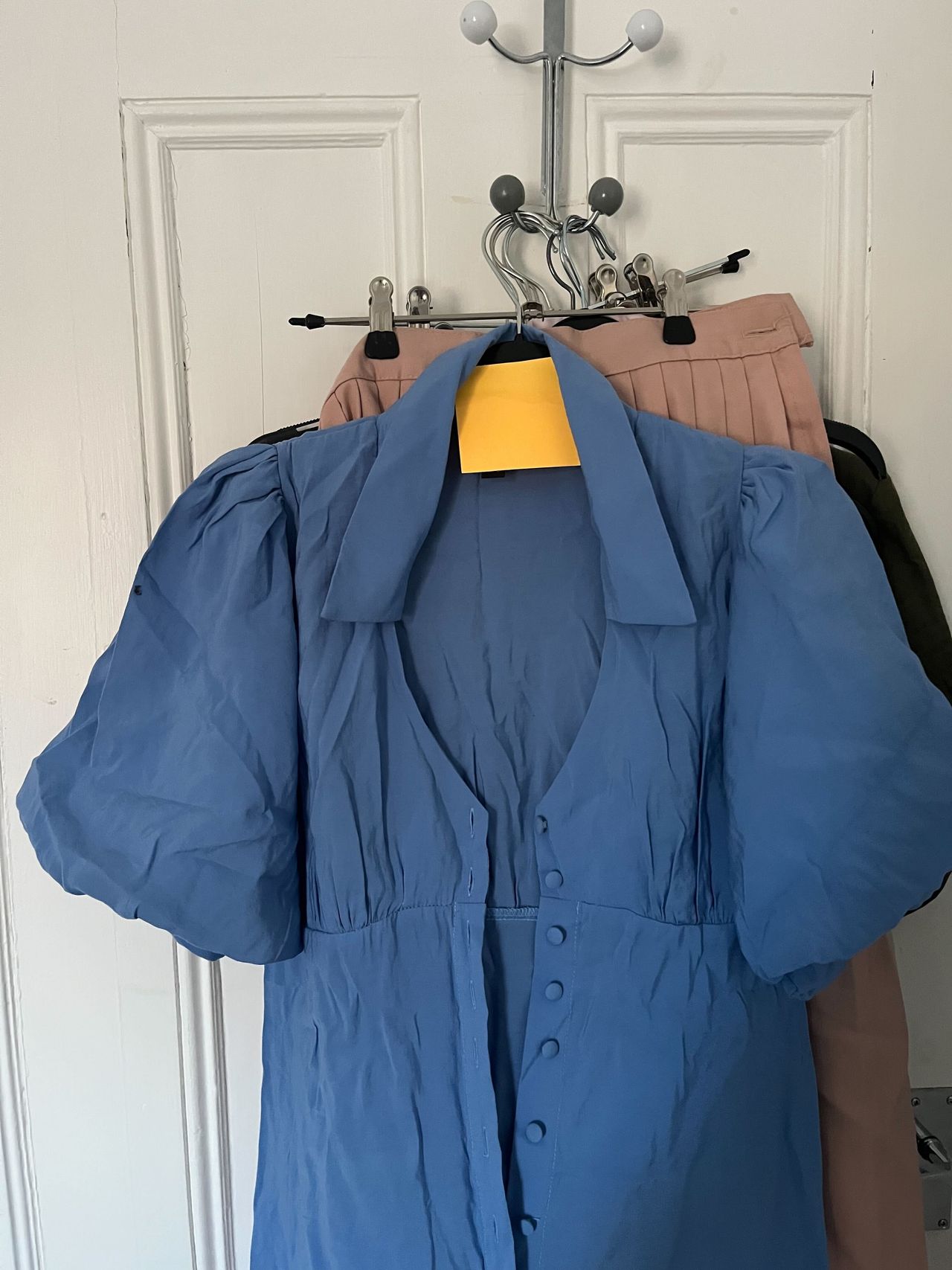 Clothes hanging on the back of a door, a blue dress at the front with an orange post it note on the hanger