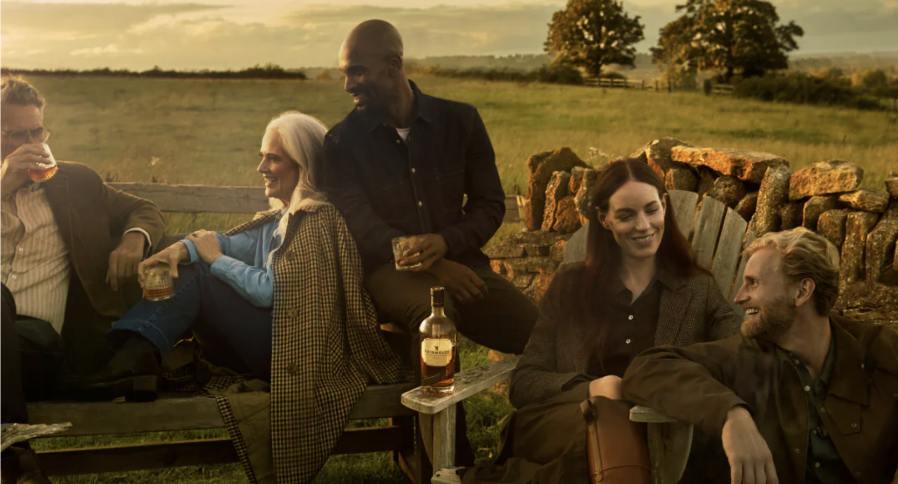 an image of five people sitting on benches at the distillery at golden hour enjoying some of the The Cotswolds Distillery whisky