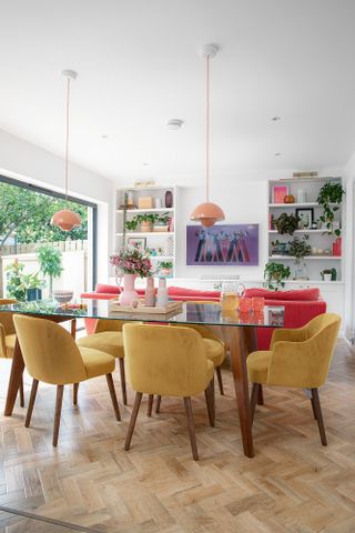 Glass-topped table and yellow dining chairs in open plan living space, with pink pendant lights overhead