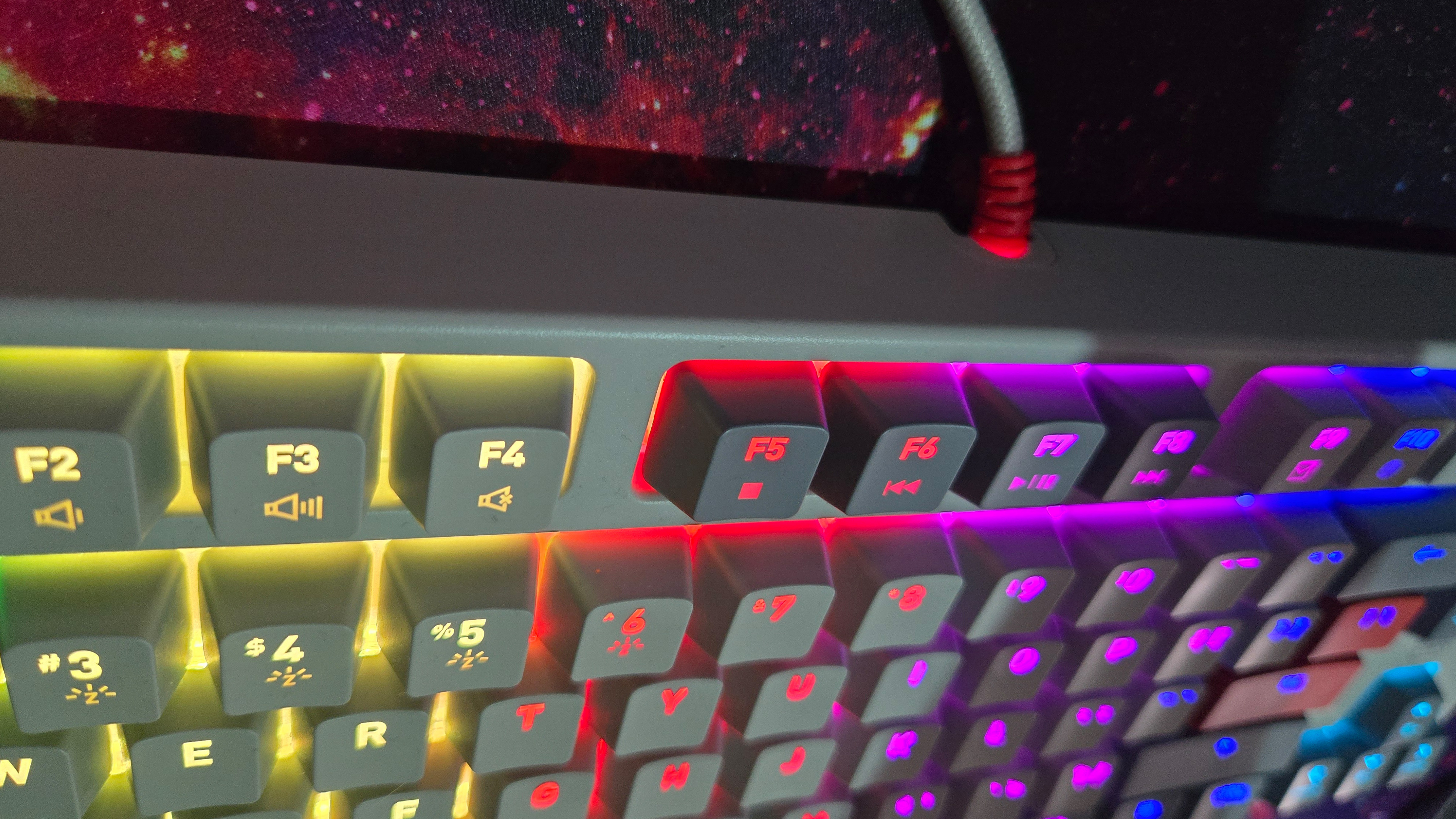 A close up of the function key row on the Cherry Xtrfy K4V2 TKL gaming keyboard. The grey braided cable with red hardware can be seen attached to the back of the keyboard. The RGB lights are on. The mousepad with the nebula design on it can be seen beneath the keyboard and braided cable.