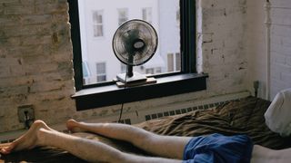 A man in blue sleep shorts sleeps on top of his duvet with a fan blowing in air from an open window
